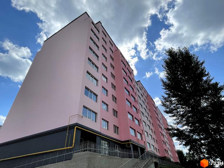 Зовнішній вигляд новобудови ЖК Світанок з рожевим фасадом та блакитним небом. Навколо будівлі видно зелені дерева та елементи прибудинкової території.