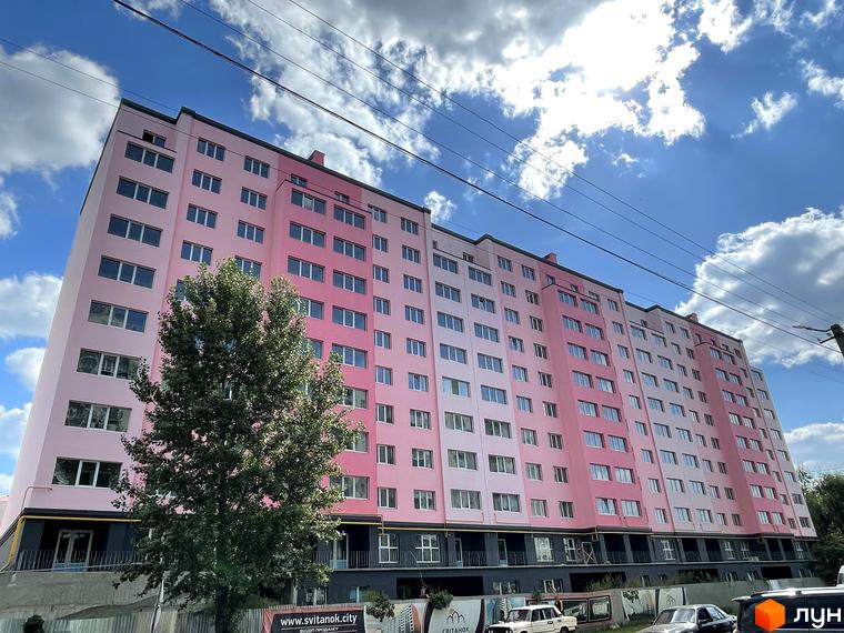 Новобудова ЖК Світанок – багатоповерховий будинок з рожевим фасадом та численними вікнами. На прибудинковій території видно дерева та паркан.