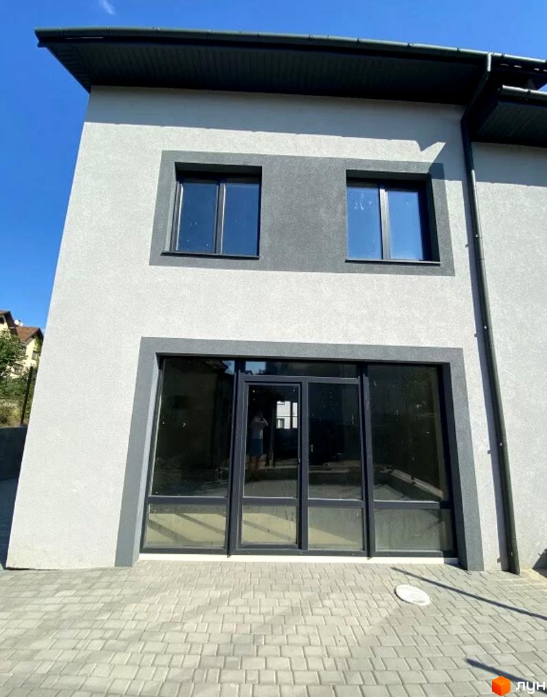 Таунхауси Hodillywood 3.0. Зовнішній вигляд будинку з великими вікнами та сірою обробкою фасаду. Поруч вимощена сіра доріжка.