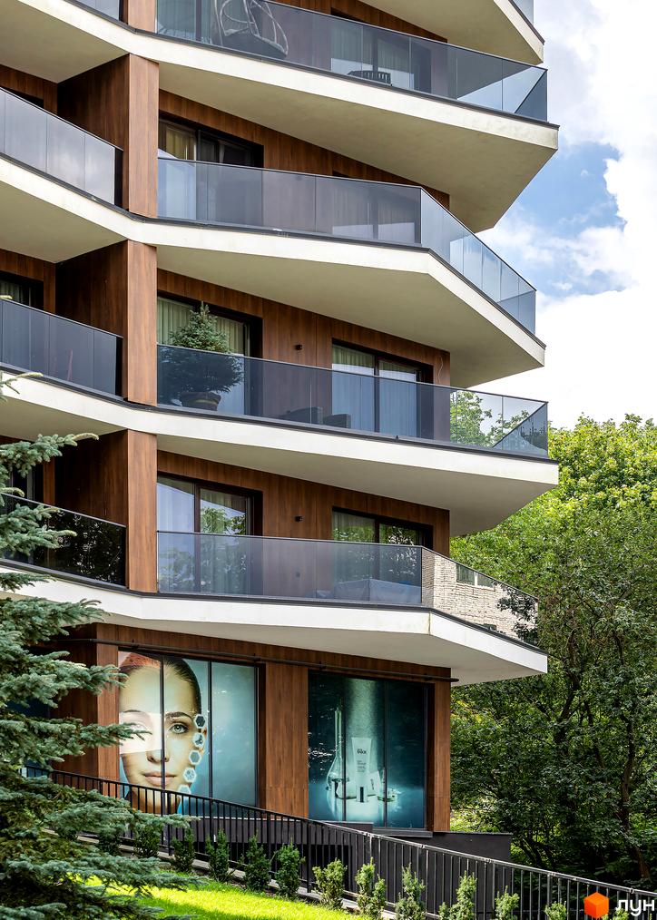 Зовнішній вигляд житлового комплексу Avalon Lux з вигнутими балконами та дерев'яними елементами фасаду. На першому поверсі вітрини з рекламою.