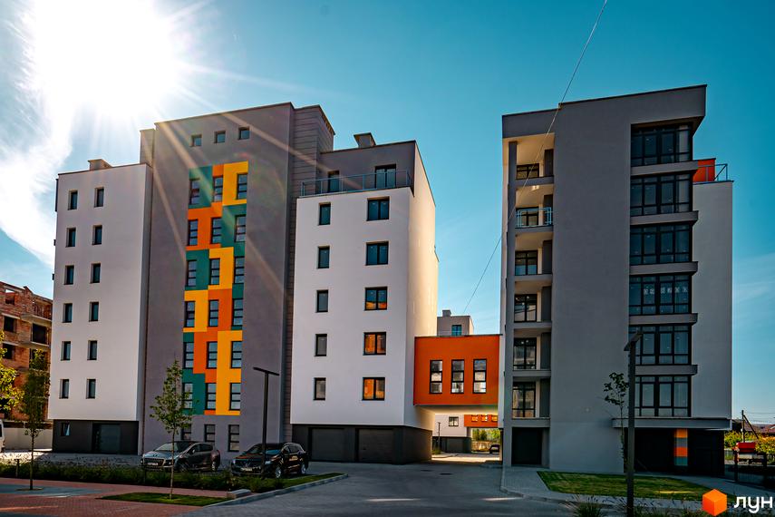 Новобудова Квартал Краківський. Будинки з білими та сірими фасадами, один з яких має кольоровий геометричний дизайн. Територія з паркінгом та деревами.