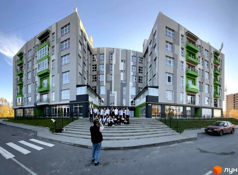 Зовнішній вигляд новобудови ЖК ЕкоПарк з фасадами сірого та зеленого кольорів. Перед будинком розташовані сходи та група людей, що фотографуються.