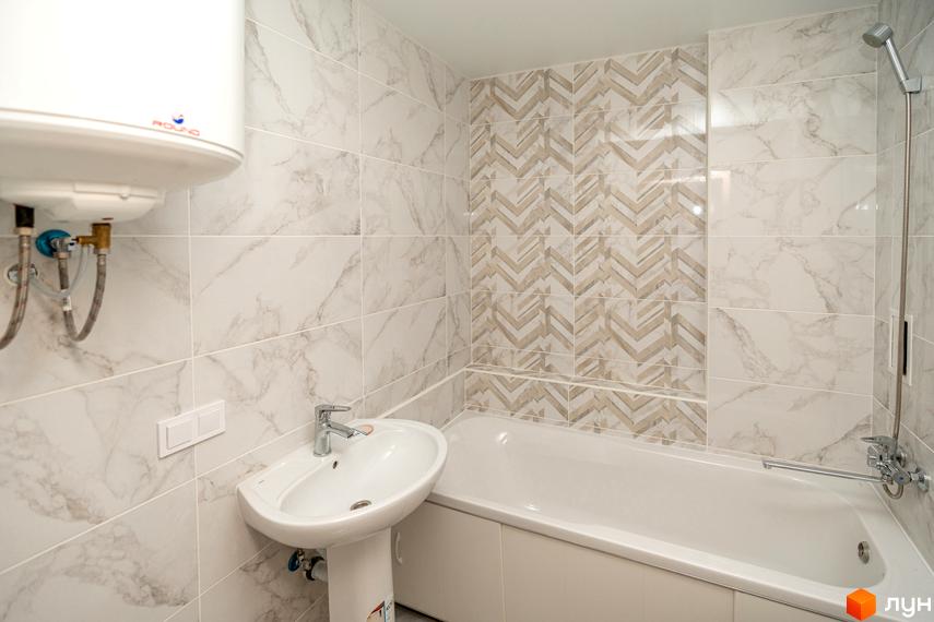Ванна кімната з білою плиткою під мармур та декоративною плиткою з геометричним візерунком. У ванній кімнаті є ванна, умивальник та водонагрівач.