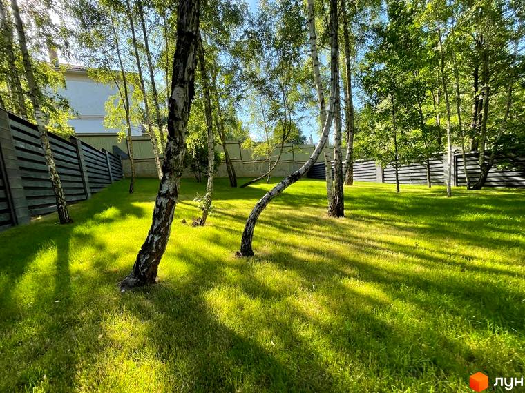 Фасад будинку Котеджного містечка Elegant House на тлі зеленого газону з березами. Територія огороджена парканом.