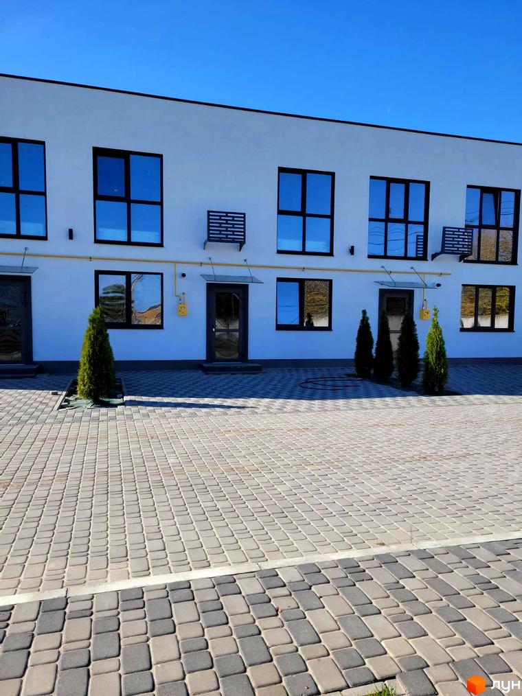 Таунхауси DREAM HOUSE з білим фасадом та великими вікнами, розташовані на вимощеній бруківкою території з зеленими насадженнями.