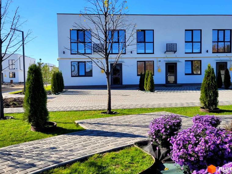 Білі двоповерхові таунхауси DREAM HOUSE з темними вікнами та вхідними дверима. Прибудинкова територія має мощені доріжки, зелені газони та кущі з фіолетовими квітами.