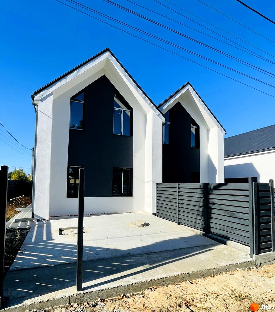 Два білих дуплекси з чорними фасадами під блакитним небом, огороджені сірим парканом. Прибудинкова територія з бетонним покриттям та зеленими насадженнями.