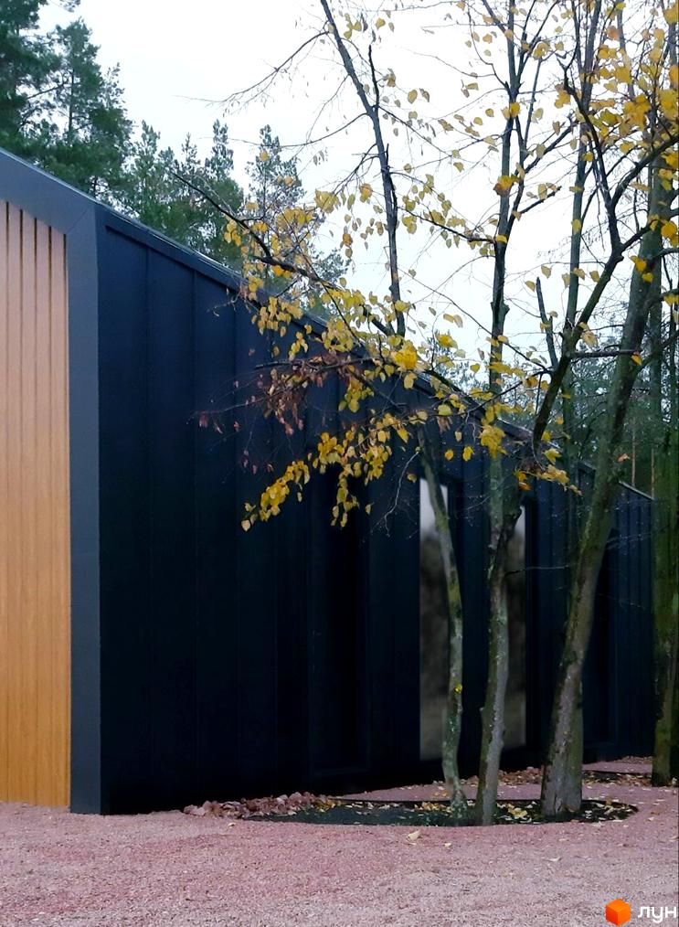 Чорний будинок з дерев'яною обробкою на тлі осінніх дерев. На передньому плані рожевий гравій.