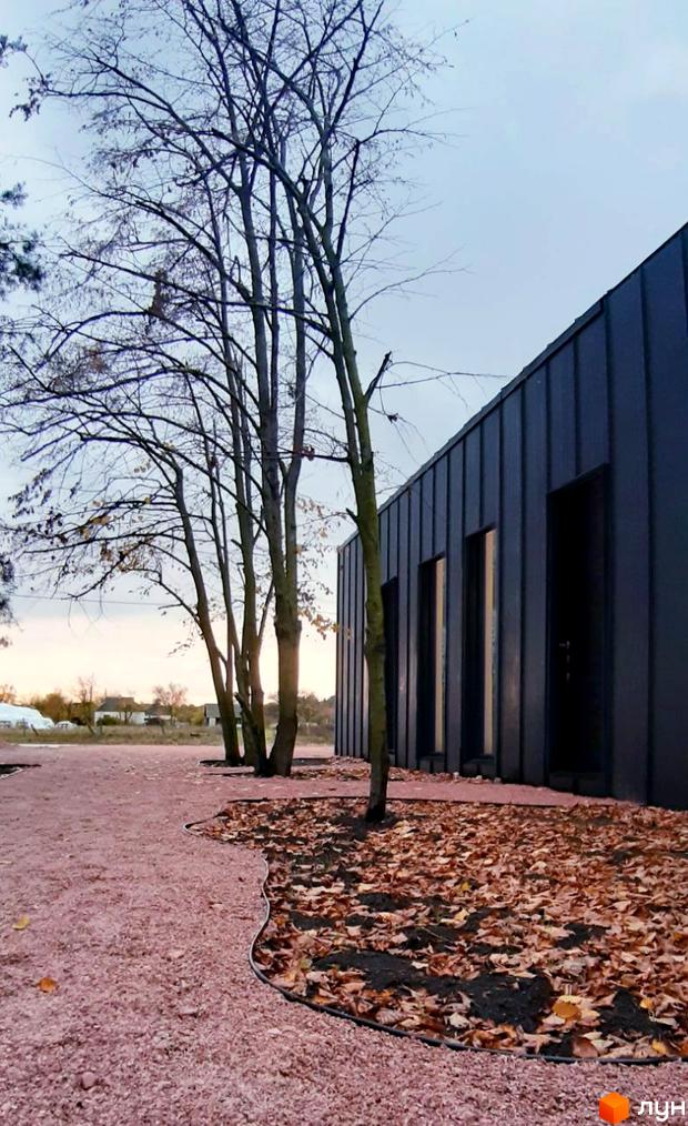 Зовнішній вигляд будинку таунхаусів Lbd1 з темним фасадом. Територія навколо будинку вкрита листям та рожевим покриттям.