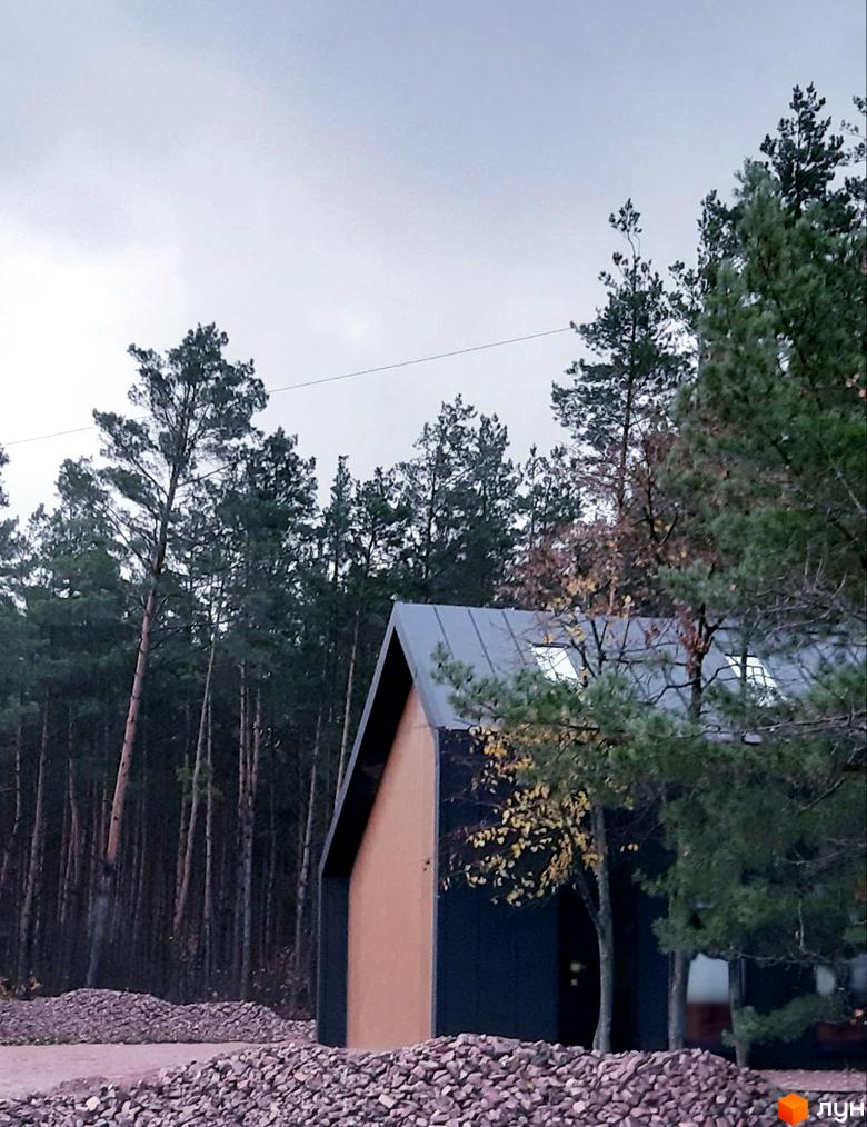 Будинок з темним фасадом та похилим дахом серед лісу. На передньому плані – кам'яниста прибудинкова територія.