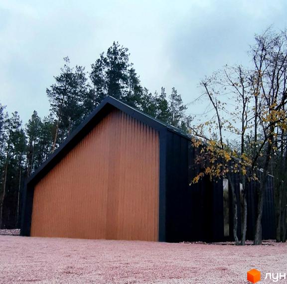 Будинок з дерев'яним фасадом у Котеджному комплексі Lbd1 на фоні лісу. Територія перед будинком вкрита рожевим щебенем.