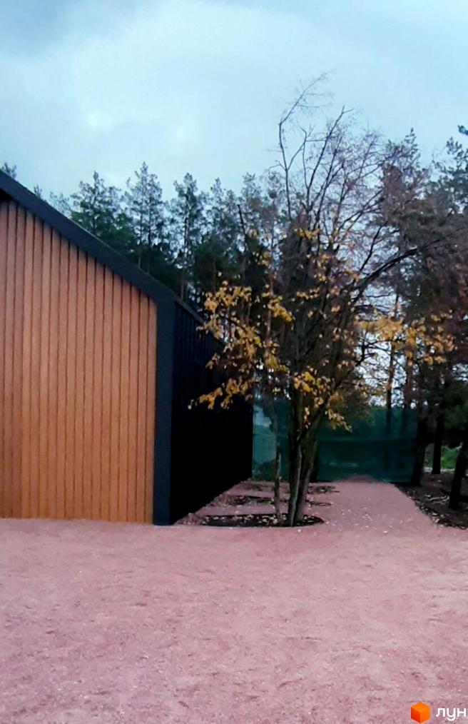 Будинок із дерев'яним фасадом у Новобудові Lbd1. Навколо будинку доріжки з рожевим покриттям та дерева.