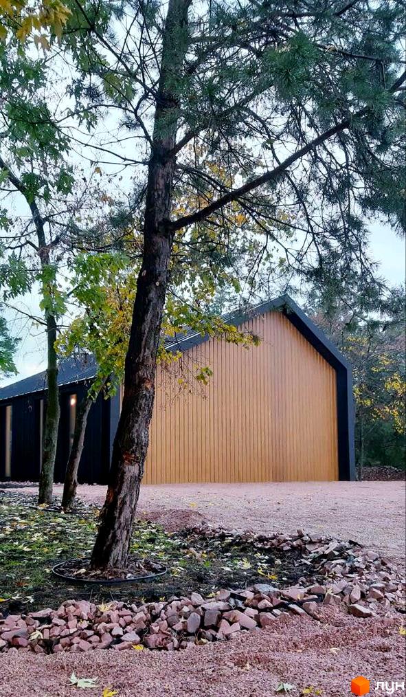 Дерев'яний будинок з чорним дахом та оздобленням. Перед будинком гравій та каміння, ростуть дерева.