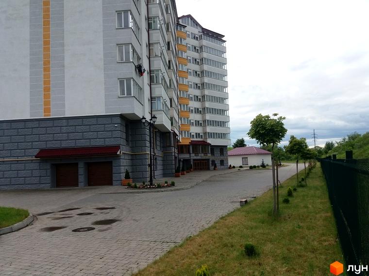 Зовнішній вигляд багатоповерхового будинку ЖК Ювілейний з сірим цоколем та гаражними воротами. Прибудинкова територія з доглянутим газоном та деревами.