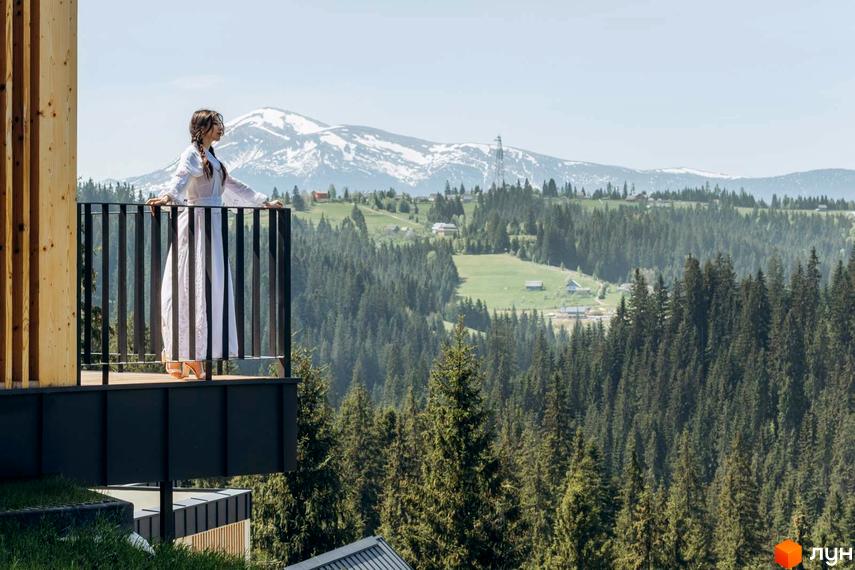Вид з балкону Котеджного містечка SKOGUR на гори та ліс. Жінка в білій сукні стоїть на балконі з видом на мальовничий пейзаж.