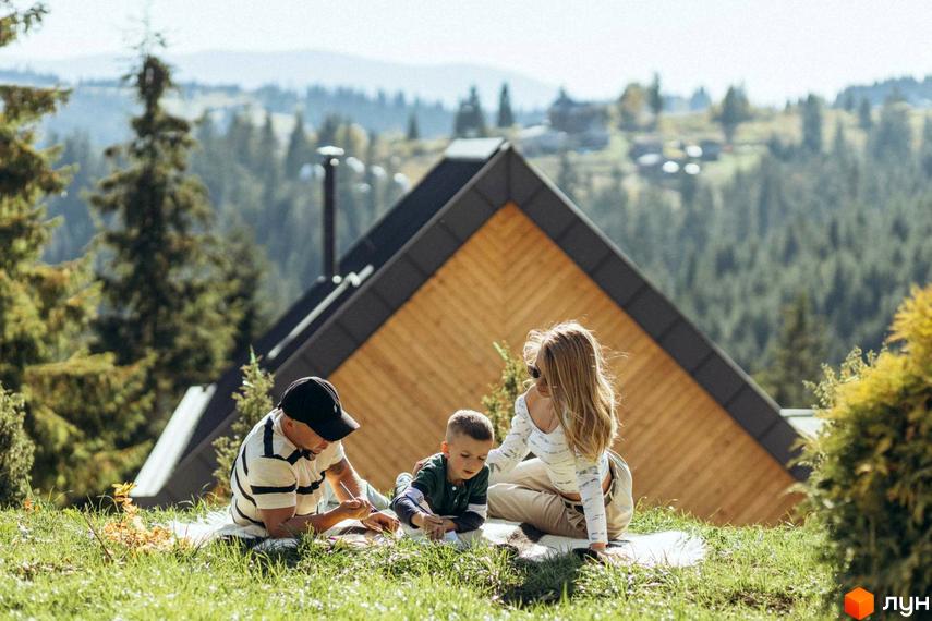 Родина відпочиває на галявині біля Котеджного містечка SKOGUR. Дерев'яний будинок з темним дахом на фоні зелених гір.