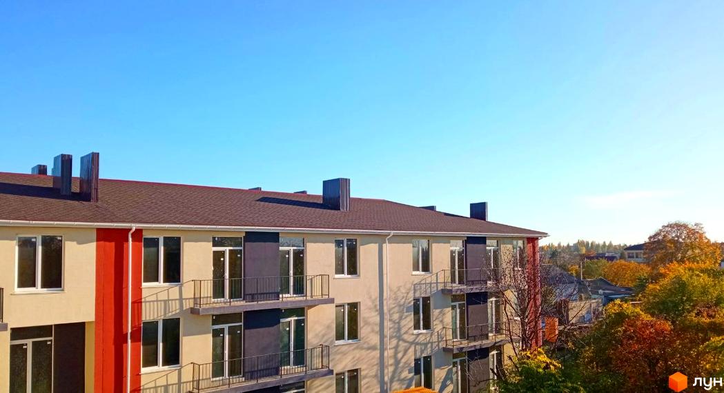 Фасад житлового комплексу Брусника з вікнами та балконами на тлі блакитного неба та осінніх дерев.