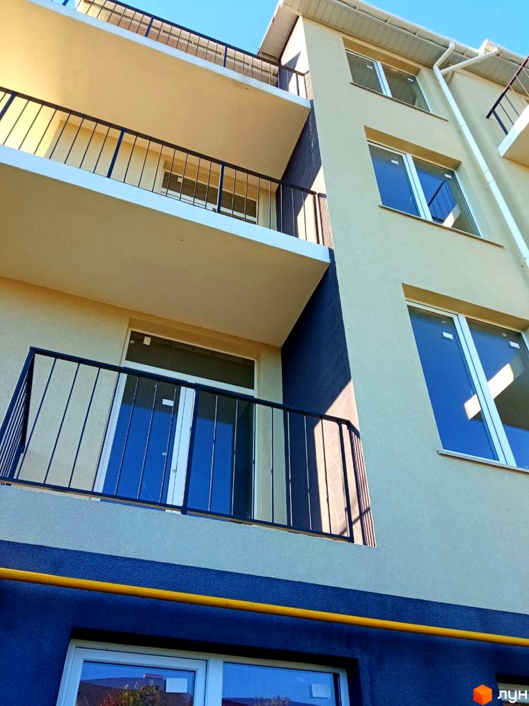 Зовнішній вигляд новобудови ЖК Брусника з балконами та вікнами на тлі блакитного неба. Будівельний проєкт з фасадом світло-бежевого кольору та синьою нижньою частиною.