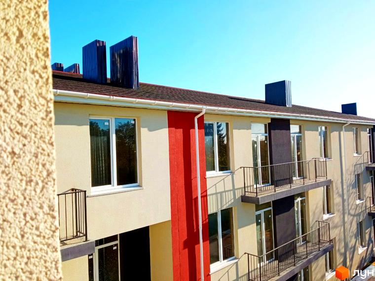 Фасад будинку Житлового комплексу Брусника: світлі стіни, червоні та темно-сірі вертикальні елементи, білі вікна, балкони з чорними перилами.