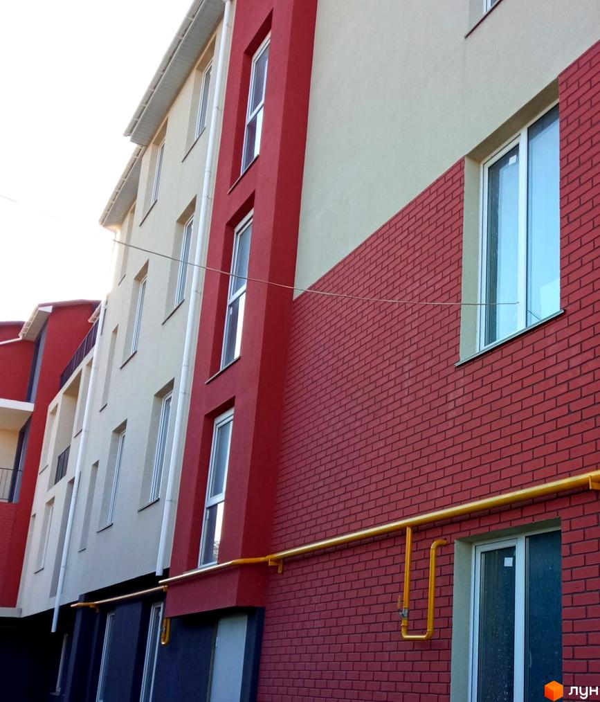 Фасад житлового комплексу Брусника з червоної цегли та бежевої штукатурки з великими вікнами. На фасаді розташовані жовті комунікаційні труби.