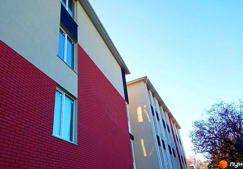 Новобудова Брусника з червоним цегляним фасадом та світлою штукатуркою, великими вікнами. На задньому плані видно дерева.