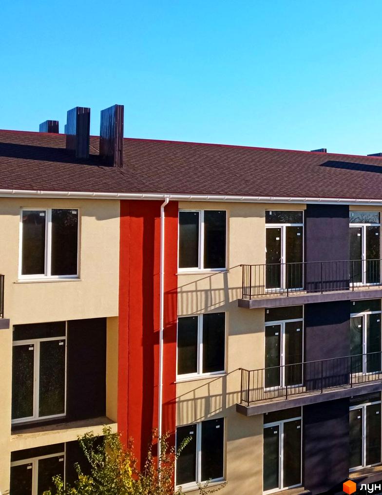 Фасад житлового комплексу Брусника з великими вікнами та балконами. Будинок має світло-бежеві та темно-сірі елементи фасаду, червону вертикальну вставку та коричневий дах.