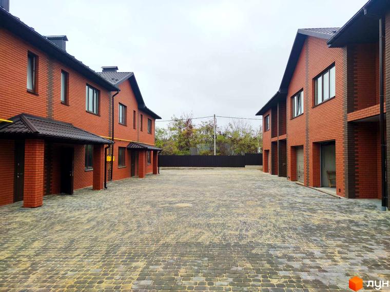 Зображення Котеджного містечка Екологічне: двоповерхові будинки з червоної цегли з темними дахами та великий двір, вимощений тротуарною плиткою.