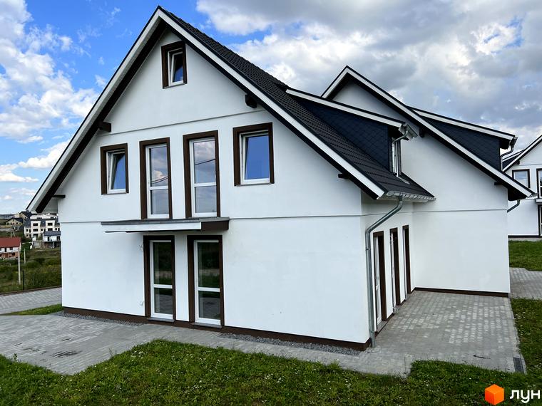 Зовнішній вигляд будинку котеджного містечка MEINDORF з білими стінами та темним дахом, розташованого на фоні хмарного неба. Поруч видно вимощену плиткою прибудинкову територію та зелену галявину.