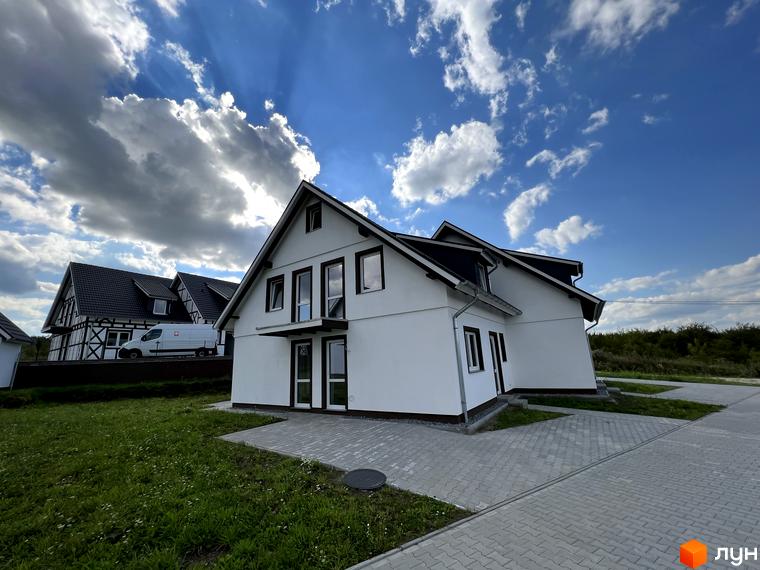 Котеджне містечко КМ MEINDORF: білий двоповерховий будинок з темним дахом та вікнами, вимощена бруківкою прибудинкова територія та зелена галявина.