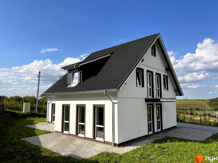 Котеджне містечко MEINDORF: білий двоповерховий будинок з чорним дахом та коричневими вікнами на тлі блакитного неба з хмарами. Навколо будинку облаштована прибудинкова територія з бруківкою та зеленою травою.