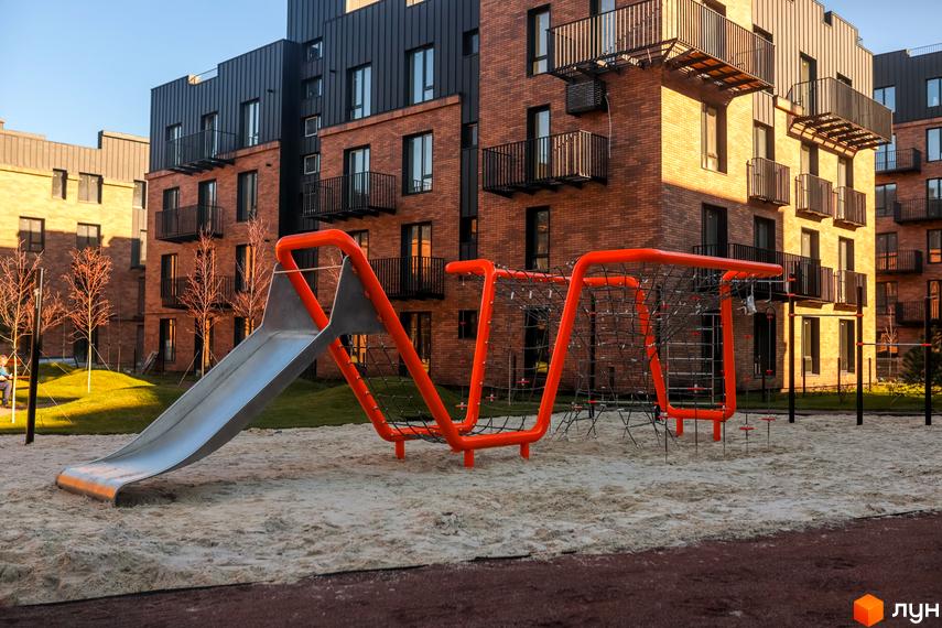 Зовнішній вигляд цегляних будинків ЖК Клубне містечко 12 з дитячим майданчиком та гіркою. Прибудинкова територія з піском та зеленню.
