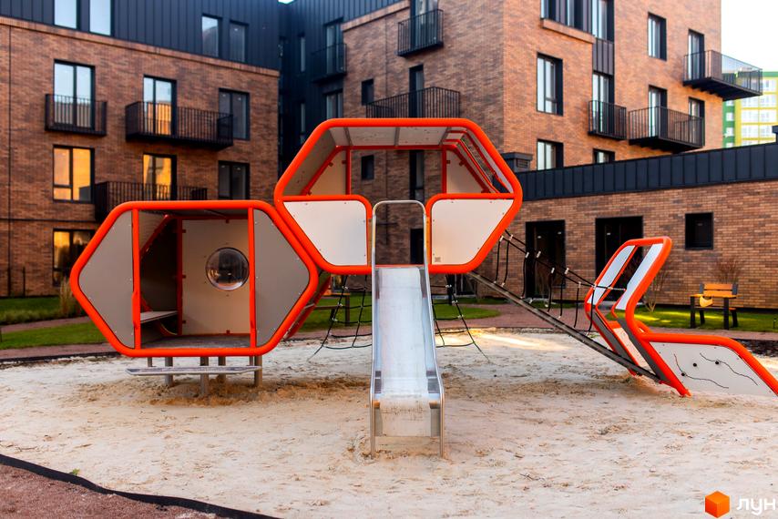 Дитячий майданчик з гіркою та ігровими елементами на території житлового комплексу Клубне містечко 12. Цегляні будинки з балконами.