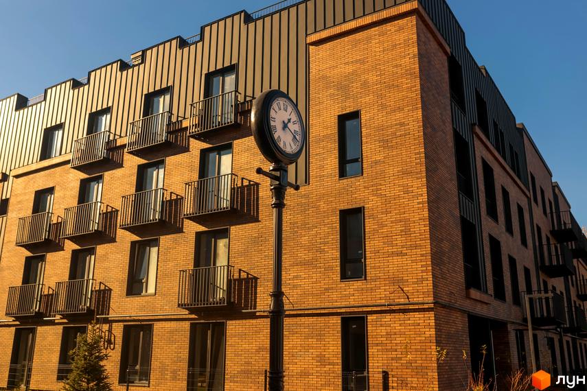 Зовнішній вигляд фасаду клубного містечка 12 з цегляним оздобленням та балконами. Навколо будівлі видно елементи прибудинкової території та вуличний годинник.