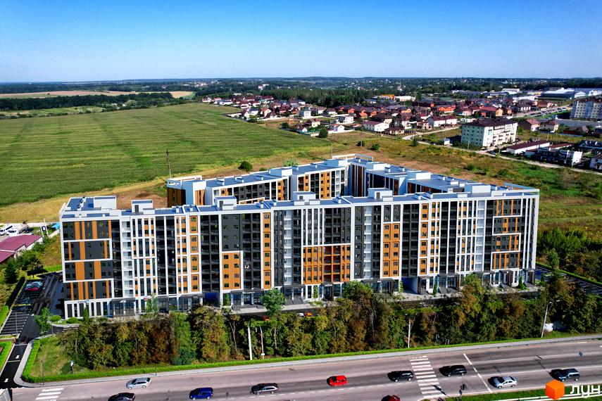 Житловий комплекс КЕЛЬЦЕ – це багатоповерхова новобудова з фасадами, оздобленими сірими та дерев'яними панелями, розташована біля зеленої зони та дороги з автомобілями.