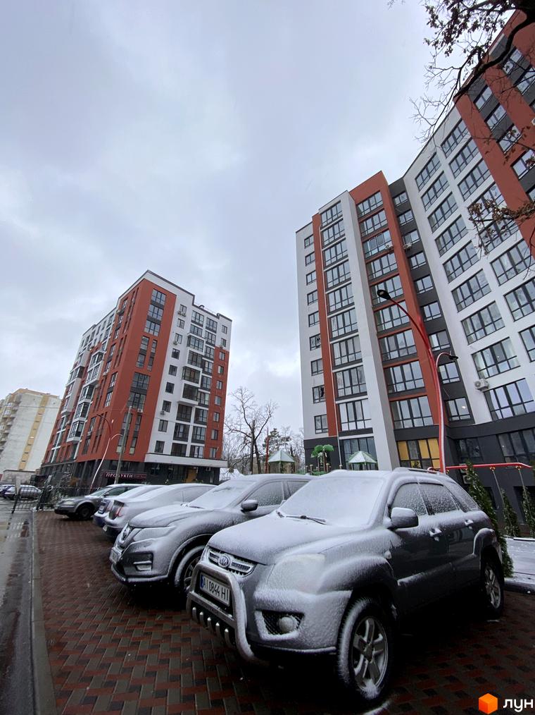 Зовнішній вигляд новобудови ЖК Green Life-3 з червоно-білими фасадами та панорамними вікнами. На прибудинковій території розташовані припарковані автомобілі та елементи дитячого майданчика.