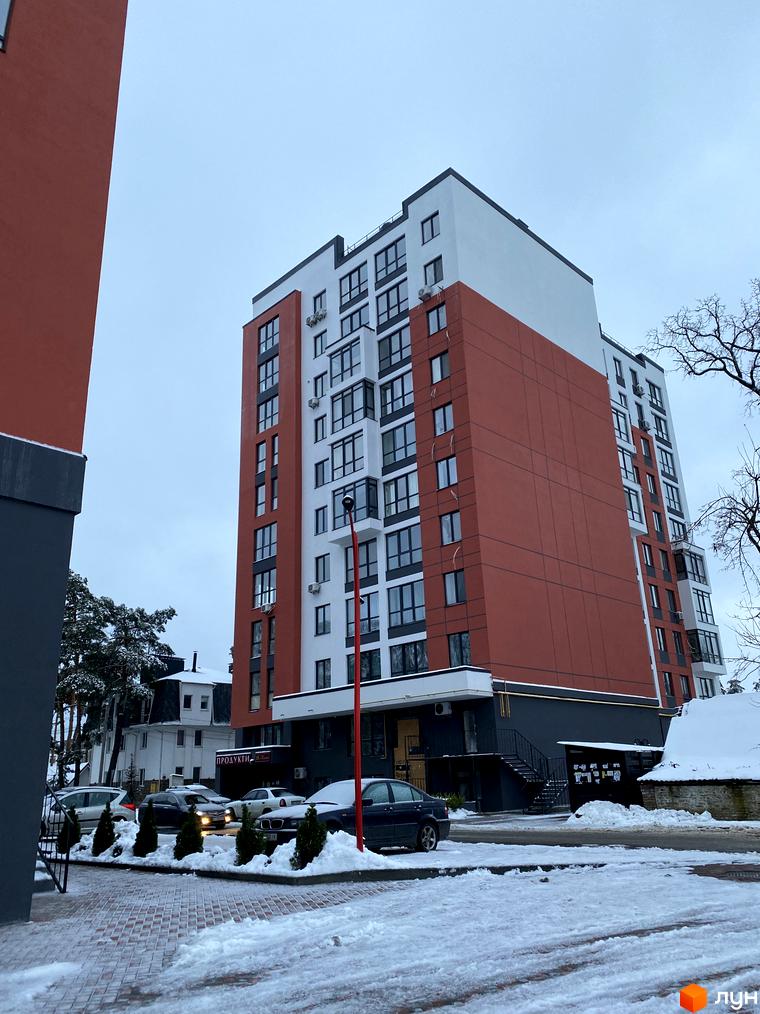 Зовнішній вигляд новобудови ЖК Green Life-3 з червоно-білим фасадом та великою кількістю вікон. Прибудинкова територія вкрита снігом, видно припарковані автомобілі та дерева.