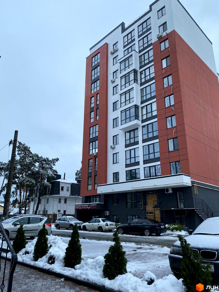 Зовнішній вигляд новобудови ЖК Green Life-3 з червоно-білим фасадом та великими вікнами. Прибудинкова територія вкрита снігом, з деревами та припаркованими автомобілями.