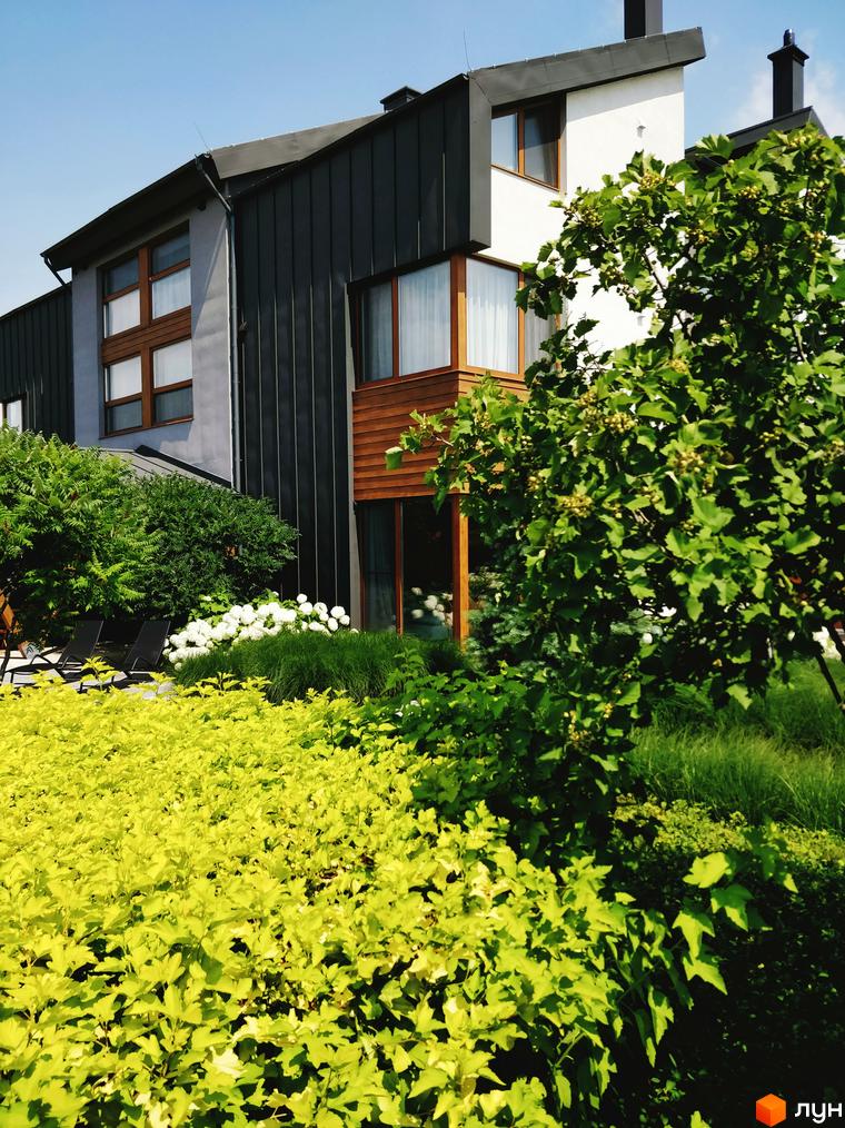 Зображення будинку з темним фасадом та дерев'яними елементами. На передньому плані зелені насадження, що створюють затишну атмосферу.