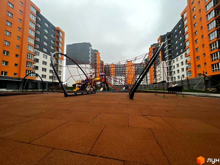 Житловий комплекс Сімейний COMFORT. Сучасний дитячий майданчик з яскравими елементами та гумовим покриттям на фоні багатоповерхових будинків з помаранчевими та сірими фасадами.