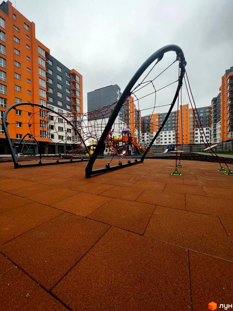 Новобудова ЖК Сімейний COMFORT з оранжевими та сірими фасадами. Дитячий майданчик з червоною сіткою та коричневим покриттям.