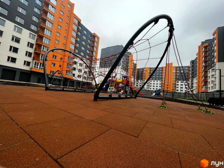 Житловий комплекс Сімейний COMFORT - сучасний дитячий майданчик біля багатоповерхових будинків з оранжевими та сірими фасадами. Територія комплексу облаштована безпечним покриттям.