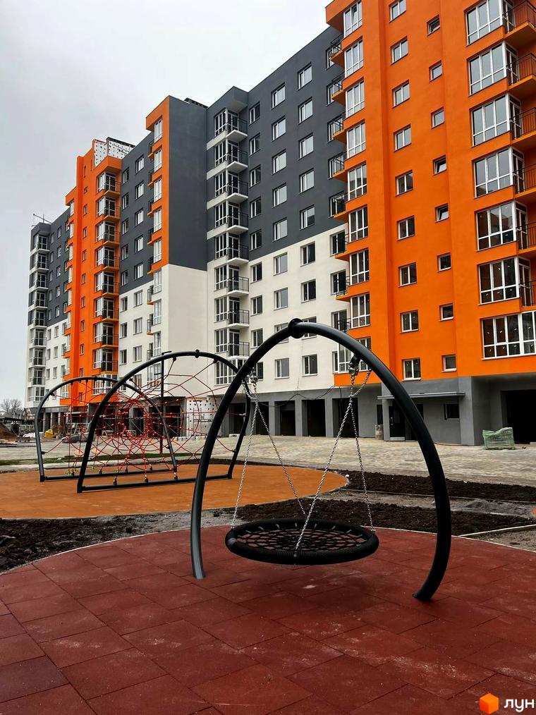 Житловий комплекс Сімейний COMFORT із яскравим фасадом в помаранчевих та сірих тонах. На прибудинковій території дитячий майданчик з гойдалками та сіткою для лазіння.