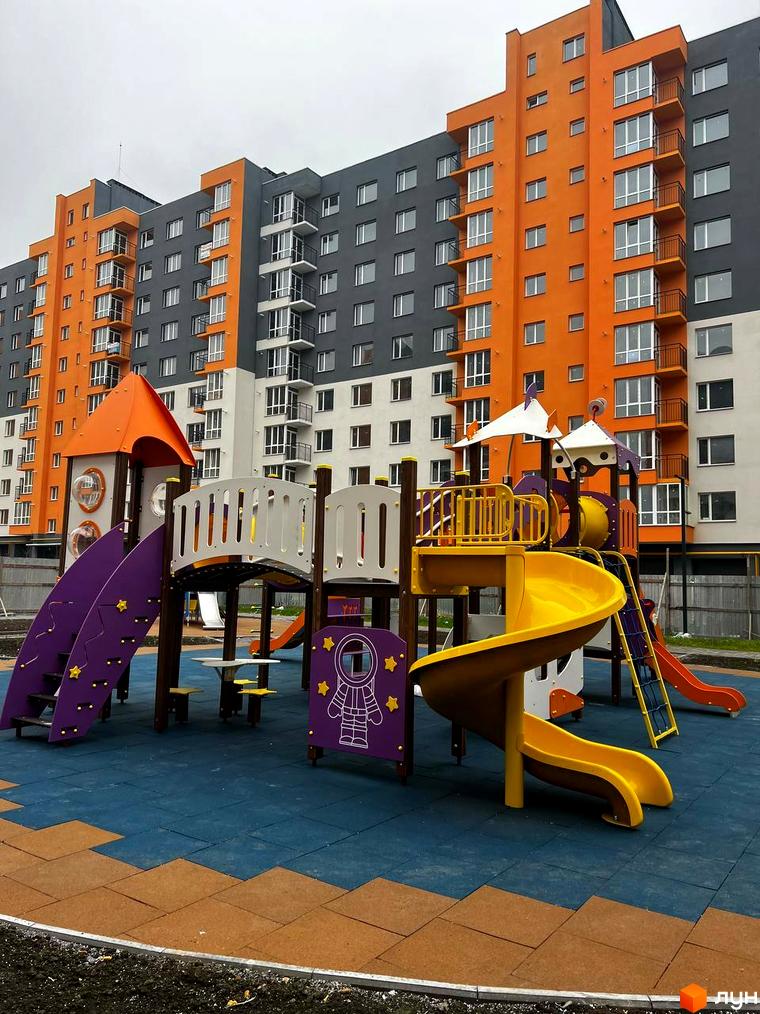 Житловий комплекс Сімейний COMFORT: яскрава фасадна частина будинку з помаранчевими та сірими секціями. Поруч дитячий майданчик з гірками та різнокольоровим покриттям.