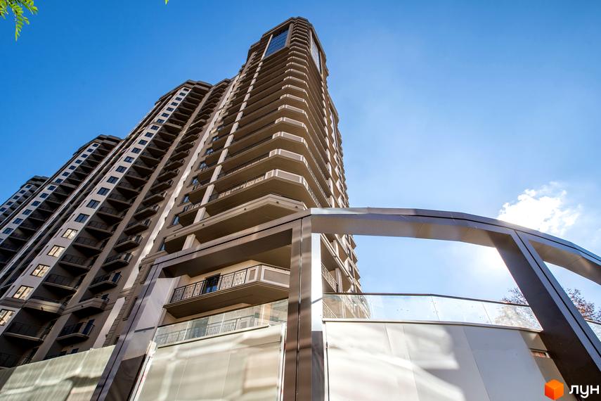 Житловий комплекс Родос: багатоповерховий будинок з балконами та світлим фасадом під блакитним небом. На передньому плані архітектурна споруда з аркою.