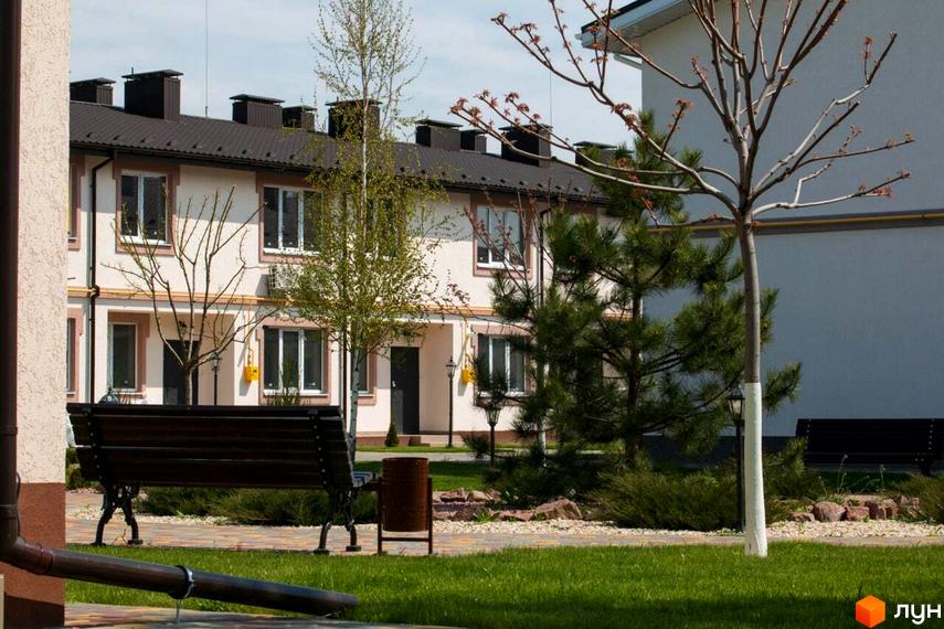 Двоповерхові будинки Котеджного містечка Паларіс зі світлими фасадами та темними дахами. На передньому плані зелений газон, дерева та лавка біля доріжки.