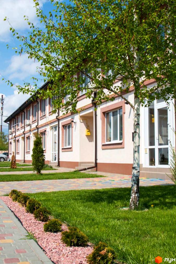 Двоповерховий будинок Котеджного містечка Паларіс зі світлими стінами та коричневими елементами. Перед будинком зелений газон, доріжка з бруківки та декоративні кущі.