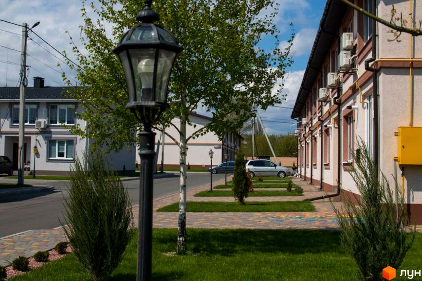 Житловий комплекс КМ Паларіс. Фасад будинку з білими стінами та коричневими віконними рамами. На прибудинковій території газон, дерева та бруківка.