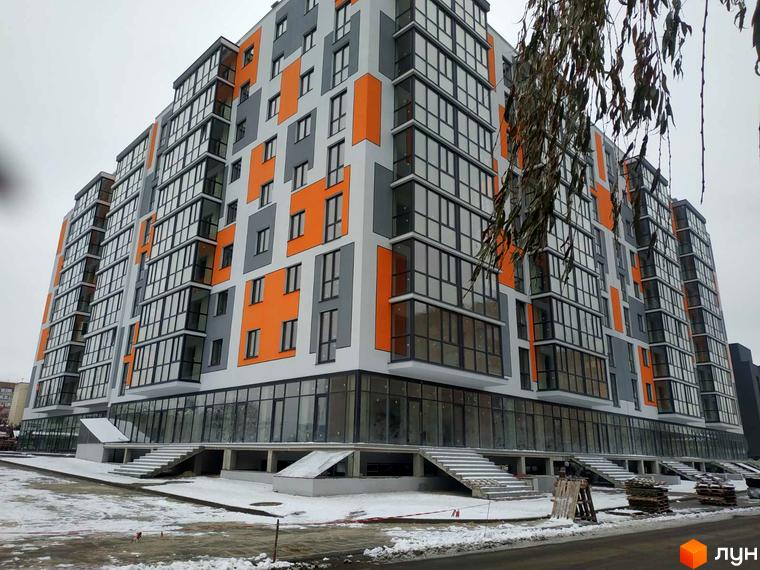 Новобудова Orange City: багатоповерхова будівля з фасадом у сірих, білих та помаранчевих кольорах. Прибудинкова територія засніжена, видно сходи до комерційних приміщень.