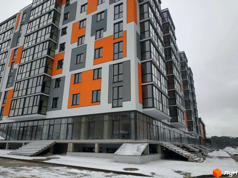 Багатоповерховий будинок Житлового комплексу Orange City з біло-сіро-помаранчевим фасадом та скляними балконами. Навколо будівлі засніжена територія з сходами.