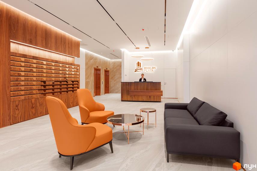 Лобі житлового комплексу Obolon Plaza з ресепшн, диваном та кріслами. Сучасний дизайн інтер'єру з дерев'яними елементами та підсвіткою.